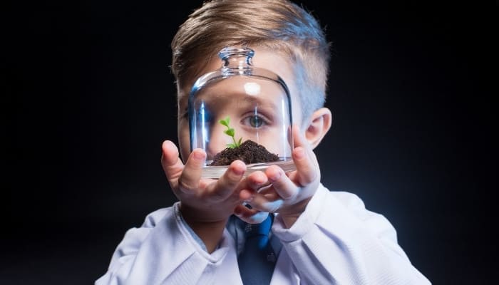 A little boy wearing a white lab coat holding a small cloche terrarium up to eye level.