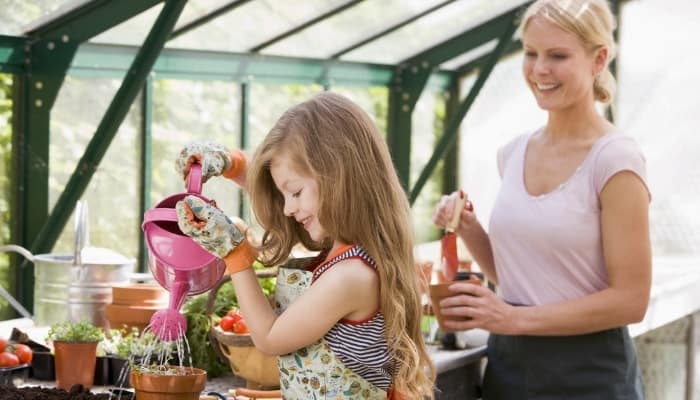 A mother watching as her daughter waters young plants in the greenhouse.