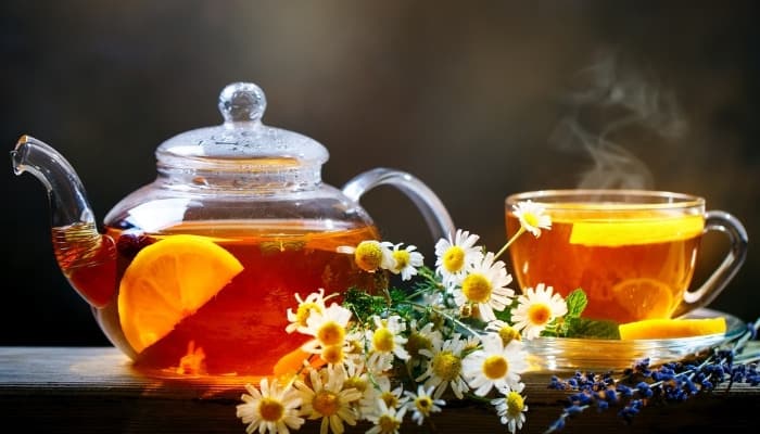 A clear tea pot and mug with freshly brewed tea placed behind cut chamomile flowers.
