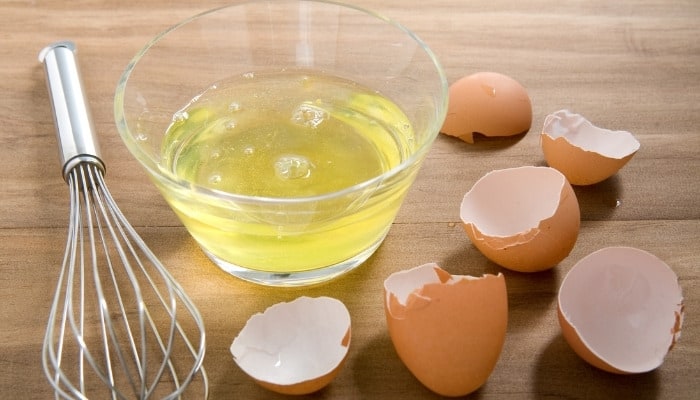 A dish of egg whites on the counter with broken egg shells and a whisk.