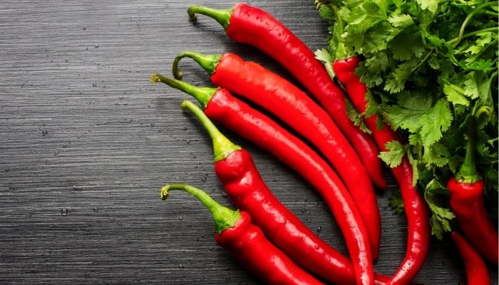 A handful of red cowhorn peppers on a wood table beside some leafy greenery.