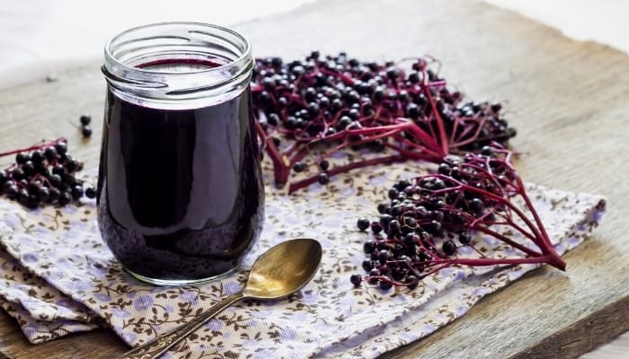 A jar of elderberry syrup on a table next to a spoon and fresh elderberries.