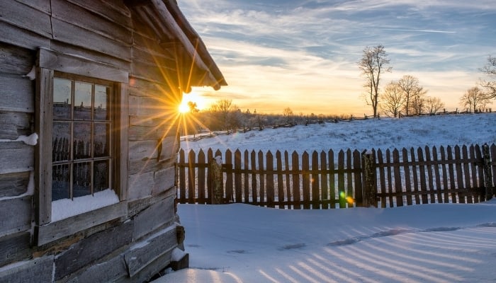 The first rays of sun peak around the corner of a rustic cabin surrounded by a picket fence.